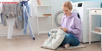 Žena sedí před pračkou se zimní bundou v ruce, na sušáku schne další oblečení