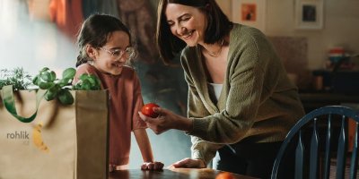 Usmívající se žena drží v ruce rajče, po jejím boku stojí usmívající se dívka, v popředí taška s nákupem