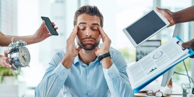 Muž v kanceláři se drží za hlavu, kolem něj ruce drží telefon, budík, tablet, složku