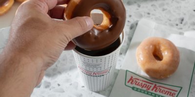 Ruka drží čokoládový donut Krispy Kreme nad kelímkem kávy