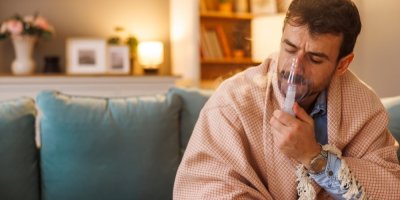 Muž přikrytý dekou sedí v obývacím pokoji a používá inhalátor 
