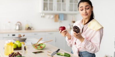 Mladá žena v kuchyni přemýšlí, zda si vezme jablko, nebo čokoládový donut