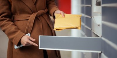 Žena v hnědém kabátu vyzvedává balíček z výdejního boxu