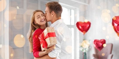 Žena dává muži dárek, ten jí dává pusu, v pozadí balónky ve tvaru srdce