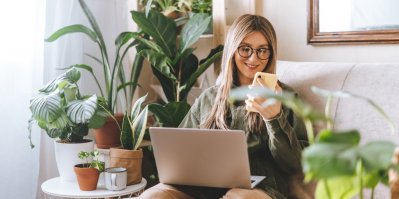 Mladý člověk sedí na gauči, na klíně má notebook, v ruce mobil a všude kolem jsou pokojové rostliny