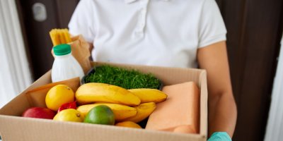 Osoba v bílém tričku drží papírovou krabici s potravinami jako banány, citrony, jablka, mléko, těstoviny, bylinky