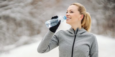 Žena v zimním oblečení stojí na zasněžené cestě a pije vodu z PET lahve