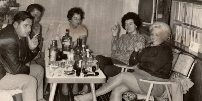 Černobílá fotografie lidí v bytě sedících kolem stolu plného alkoholu