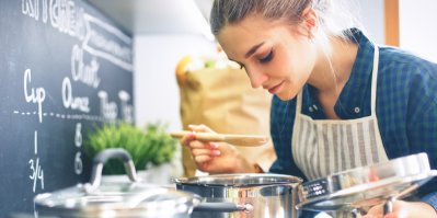 Žena v kuchyni vaří polévku v hrnci, v rukou drží pokličku a naběračku