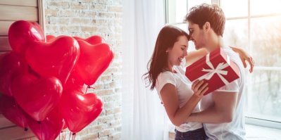 Mladý muž a žena se objímají, žena má v ruce dárek, u stěny jsou balónky ve tvaru srdce