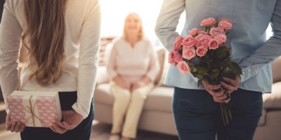 Dívka schovává dárkově zabalenou krabičku za zády, žena vedle ní má za zády kytici růží, čelem k nim sedí jejich maminka/babička na gauči