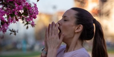 Žena si zakrývá obličej rukama při kýchnutí, za ní jsou rozkvetlé stromy
