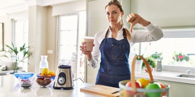 Žena v zástěře stojí v kuchyni u mixéru, v ruce má smoothie a druhou rukou ukazuje palec dolů