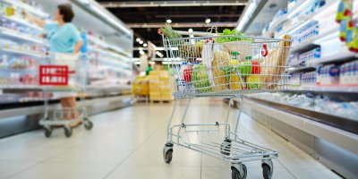 Nákupní vozík plný potravin v uličce supermarketu, v pozadí si žena vybírá zboží z regálu