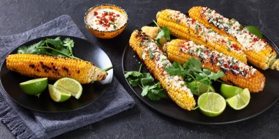 Čtyři kukuřice elotes na talíři, jedna opečená kukuřice na druhém talíři, limetky, bylinky, majonéza