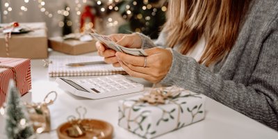 Žena ve svetru sedí mezi vánoční výzdobou a dárky a počítá peníze na kalkulačce