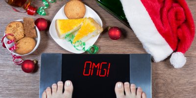 Člověk stojí na váze, která místo čísla ukazuje nápis OMG!, kolem je vánoční čepice, ozdoby a sladké pečivo