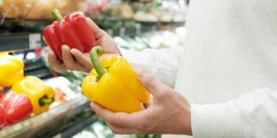 Člověk v obchodě drží jednu červenou a jednu žlutou papriku