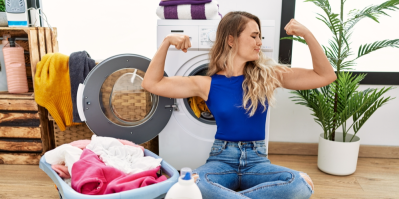 Žena sedí u pračky s košem na prádlo a ukazuje bicepsy