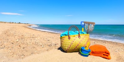 Žlutá taška na pláži, vedle ní modrý kyblíček na písek s lopatkou a červená osuška.