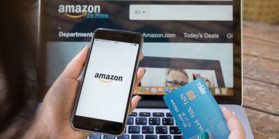 Na notebooku jsou stránky e-shopu Amazon, člověk se dívá na mobil a drží platební kartu