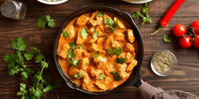 Butter chicken s rýží, servírované v hnědém talíři, v pozadí chléb naan a misky s kořením