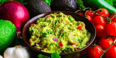 Miska s guacamole, kolem ní avokádo, česnek, rajčata a červená cibule