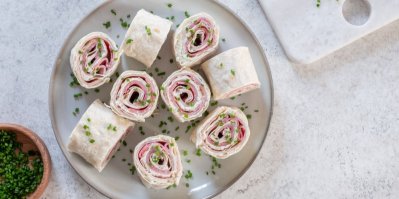 4 tortilla rolády, servírované na hnědém prkénku, vedle miska se salátem a rajčaty