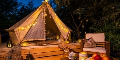 Glampingový stan vybavený nábytkem, ověšený světýlky. Před stanem je pohodlné posezení s křesílkem a stolečkem, na kterém stojí lampa, mísa s ovocem, čajová konvice a váza