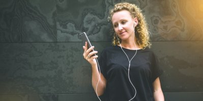 Žena s kudrnatými vlasy má sluchátka a dívá se do mobilu