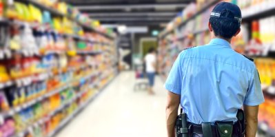 Muž v uniformě ochranky sleduje v obchodě zákazníka mezi regály.