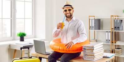 Zaměstnanec sedí na stole v kanceláři, má na sobě sluneční brýle, nafukovací kruh a pije limonádu