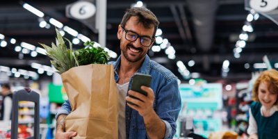 Usměvavý muž se dívá do telefonu na nákupech potravin v obchodě