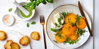 Plátky kaki, rukola a burrata na talíři, bazalkové pesto a pečené plátky rohlíku