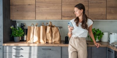 Žena s chytrým telefonem v ruce u kuchyňské linky, na které stojí papírové tašky s nákupem