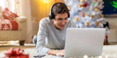 Teenager leží na břiše před vánočním stromkem, na hlavě má sluchátka a před sebou notebook