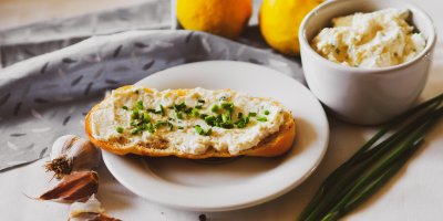 Krajíc chleba s česnekovou pomazánkou na bílém talíři, kolem stroužky česneku