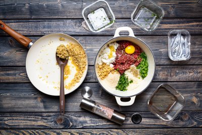 Mleté maso, vejce, cibule a další suroviny v hrnci, vedle cibule na pánvi.