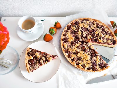 Třešňový koláč s tvarohem naservírovaný na talíři, vedle zbytek koláče a šálek kávy