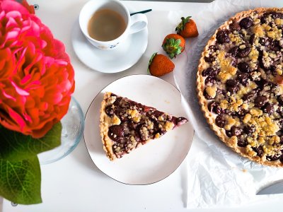 Třešňový koláč s tvarohem naservírovaný na talíři, vedle zbytek koláče a šálek kávy