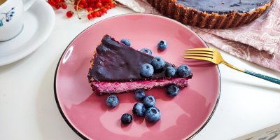 Borůvkový cheesecake na růžovém talíři, ozdobený borůvkami, v pozadí zbytek dortu a káva