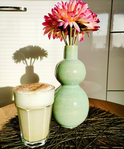 Matcha latte ve sklenici se skořicí, vedle sklenice je postavena váza s květinou