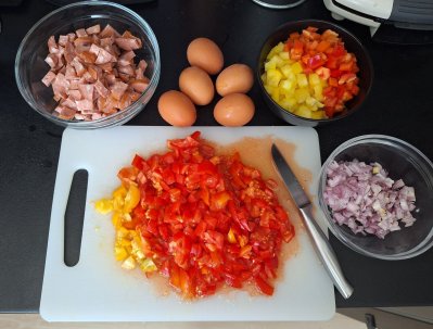 Pokrájená rajčata na bílém prkénku, pokrájená cibule, paprika, špekáčky v misce, vejce