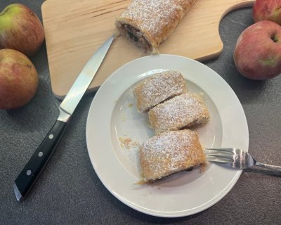 Nakrájený jablečný štrúdl na bílém talířku, vedle prkénko, jablka, nůž a vidlička