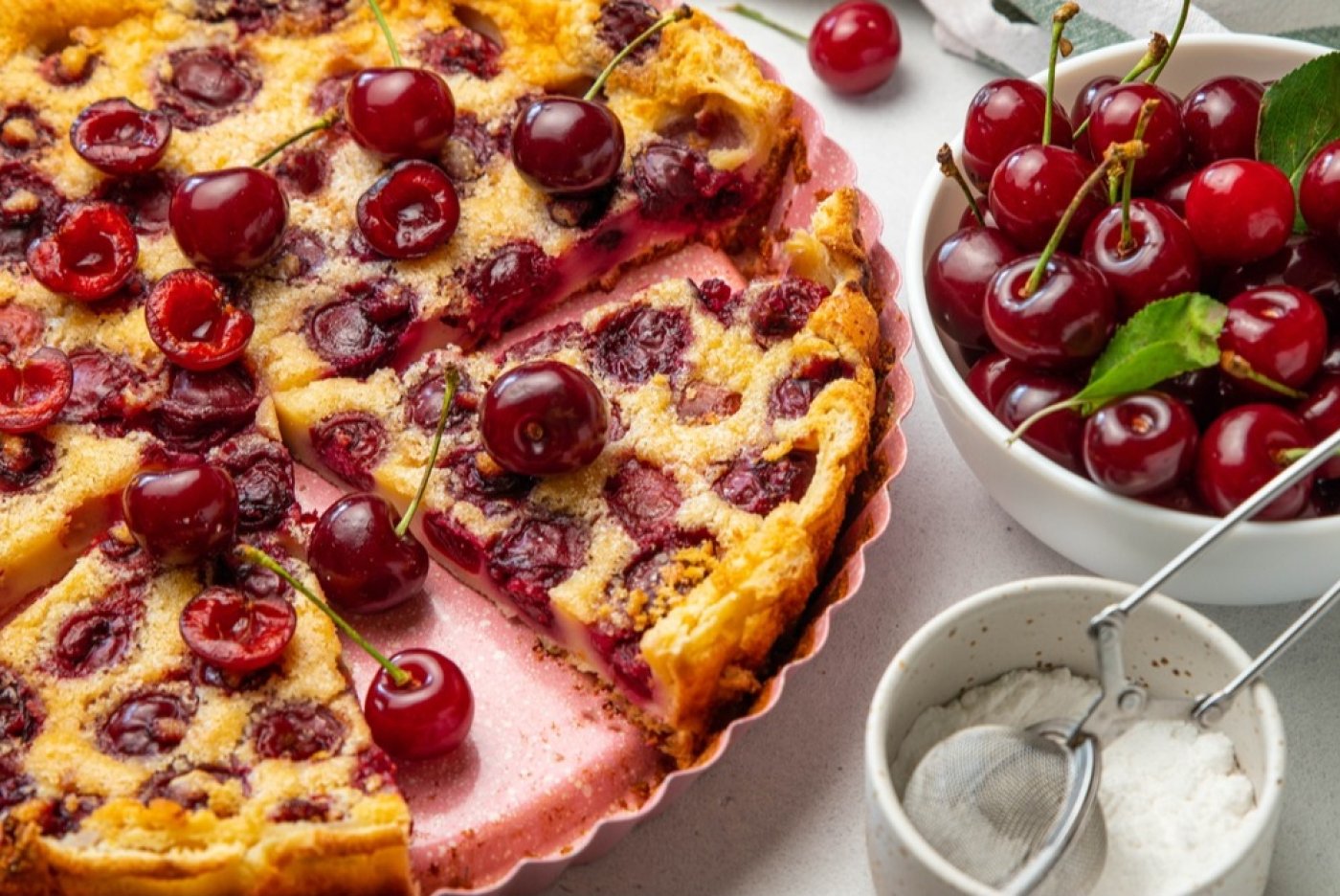 Třešňový koláč clafoutis v koláčové formě, vedle miska s třešněmi a miska s cukrem