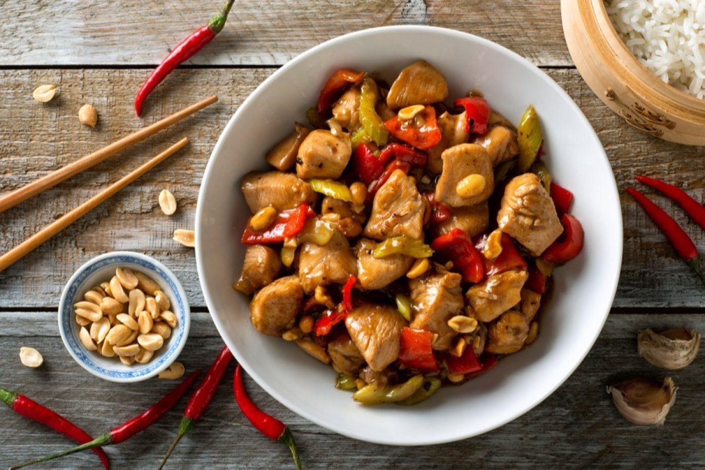 Kuřecí kung pao v bílé misce, na dřevěném stole, vedle stroužky česneku, chilli papričky, mistička s arašídy, jídelní hůlky a miska s rýží