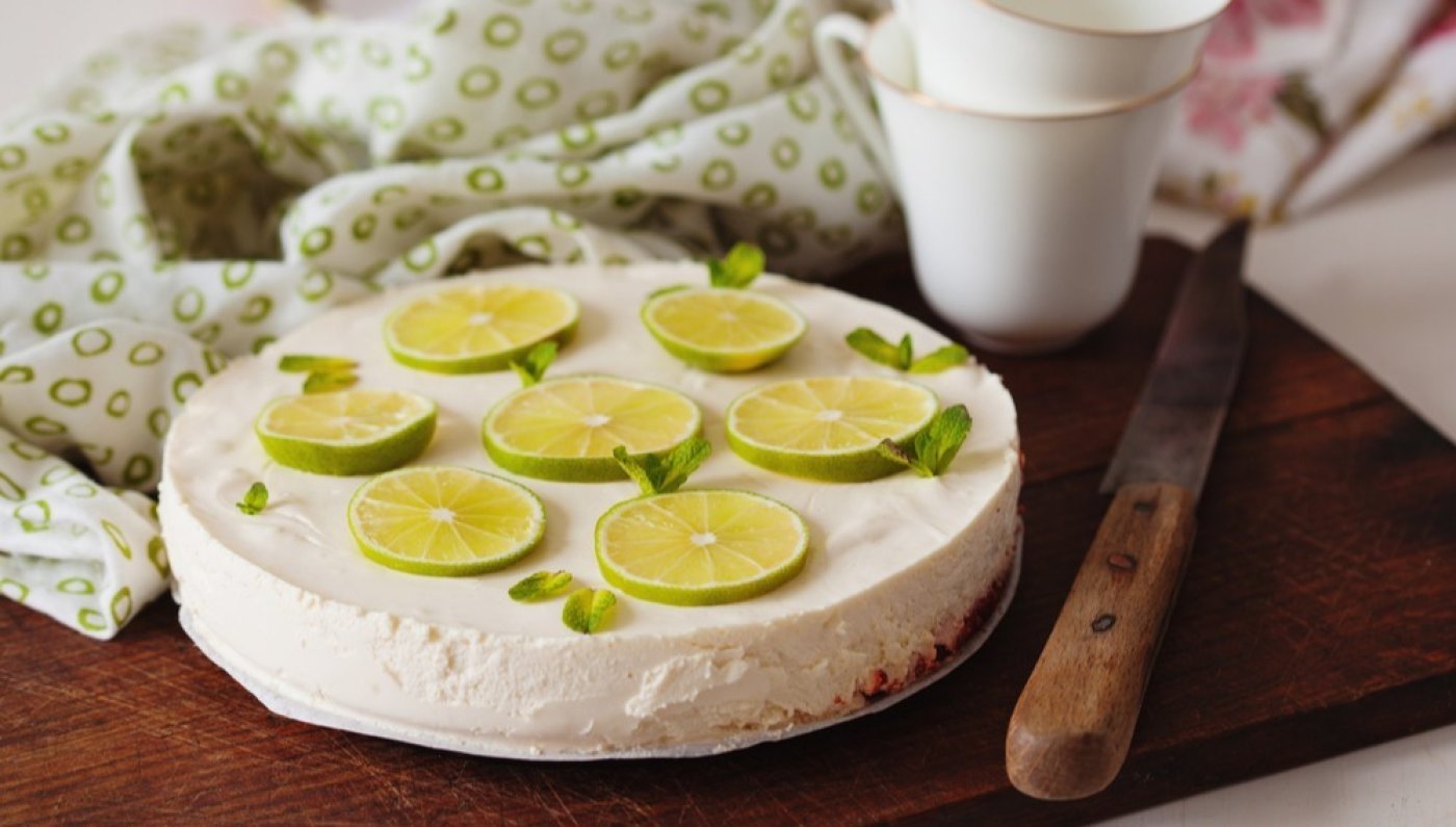 Nepečený limetkový cheesecake ozdobený kolečky limetky, na dřevěném prkénku, v pozadí bílo-zelená utěrka