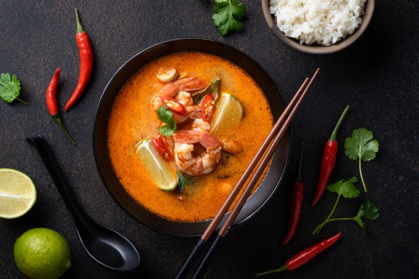 Thajská polévka Tom Yum servírovaná v černé polévkové misce, vedle misky lžíce, rozkrojená limeta a chilli papričky